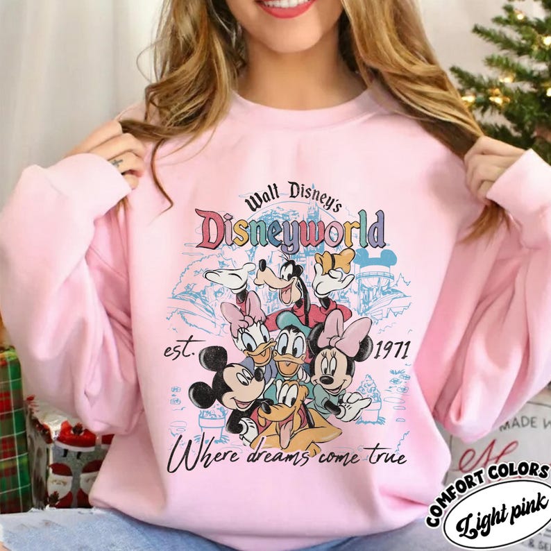 May include: Light pink sweatshirt featuring a Disneyworld graphic with Mickey Mouse, Minnie Mouse, Donald Duck, Daisy Duck, Goofy, and Pluto. The design includes the text "Walt Disney's Disneyworld", "est. 1971", and "Where dreams come true".