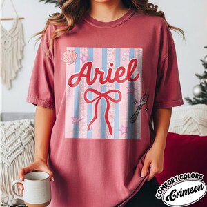 May include: Crimson-colored t-shirt featuring a graphic design with the name "Ariel" in red script. The design includes a bow, stars, a seashell, and a fork on a striped background. The shirt is a Comfort Colors brand.