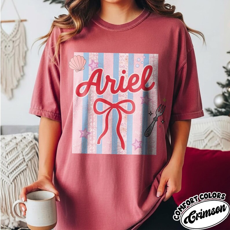 May include: Crimson-colored t-shirt featuring a graphic design with the name "Ariel" in red script. The design includes a bow, stars, a seashell, and a fork on a striped background. The shirt is a Comfort Colors brand.