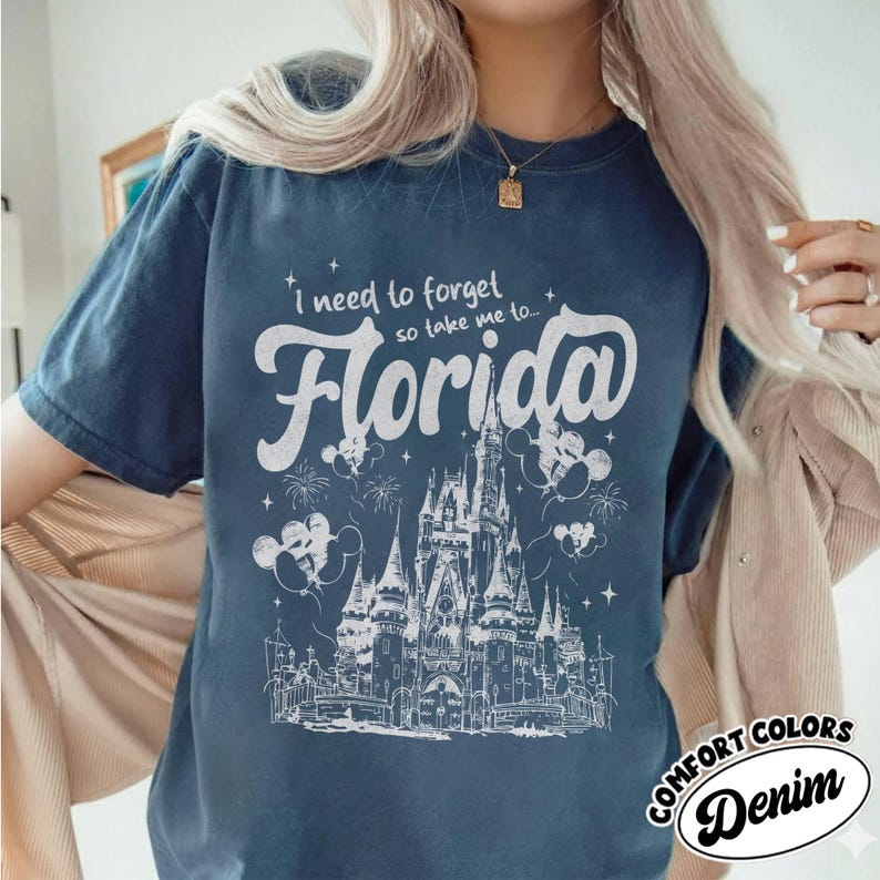May include: Navy blue t-shirt featuring a white graphic of a castle, fireworks, and Mickey Mouse balloons. The text reads "I need to forget so take me to... Florida". The shirt also has the words "Comfort Colors Denim".