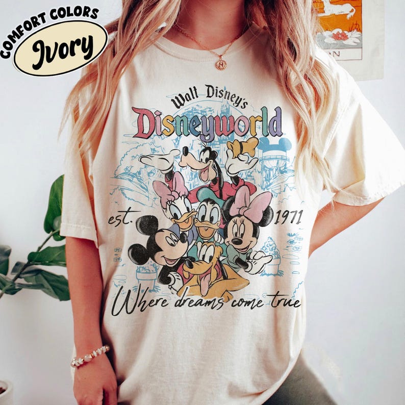 May include: Ivory-colored t-shirt featuring a colorful graphic of Disney characters, including Mickey Mouse, Minnie Mouse, Donald Duck, and Goofy. The design includes the text "Walt Disney's Disneyworld" and "Where dreams come true", with the year "1971".