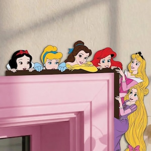 May include: A decorative wall decal featuring six illustrated Disney princesses: Snow White, Cinderella, Belle, Ariel, Aurora, and Rapunzel. The characters are arranged to appear as if they are peeking over the top and side of a door frame.