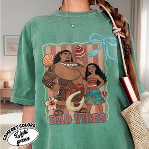 May include: Light green t-shirt with a graphic featuring two cartoon characters and the text "DAD VIBES." The graphic includes a man and a woman, along with decorative elements like seashells and a flower. The shirt also has the words "COMFORT COLORS" and "Light green."