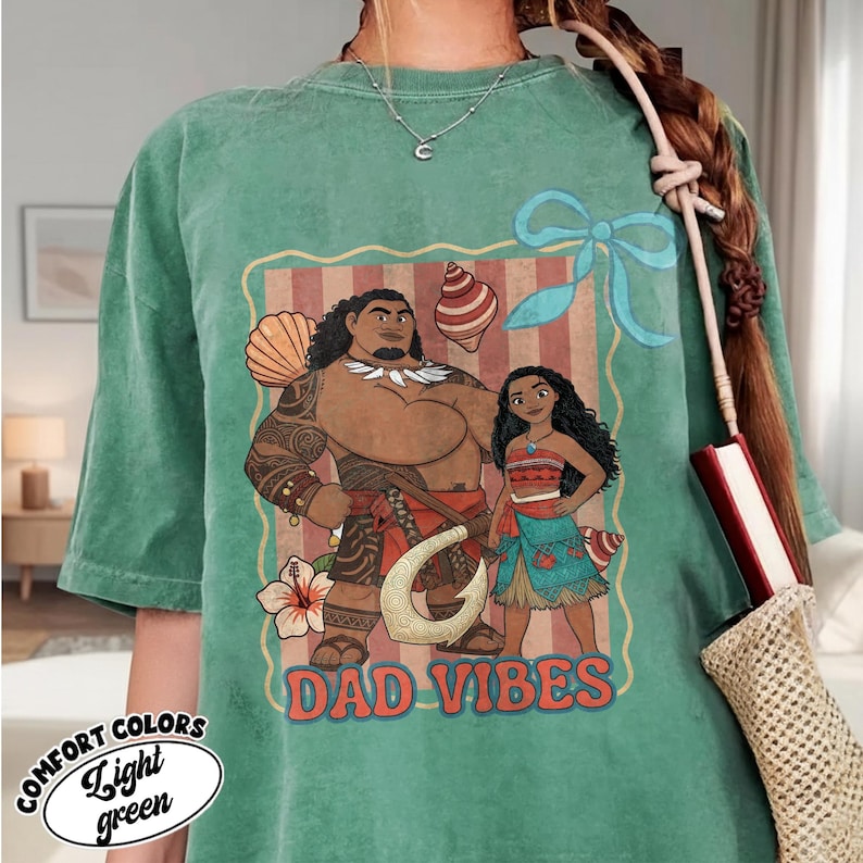 May include: Light green t-shirt with a graphic featuring two cartoon characters and the text "DAD VIBES." The graphic includes a man and a woman, along with decorative elements like seashells and a flower. The shirt also has the words "COMFORT COLORS" and "Light green."