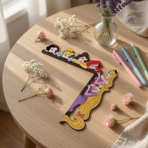 May include: An L-shaped decorative frame featuring colorful illustrations of six animated princesses, including Snow White, Cinderella, Belle, Ariel, Aurora, and Rapunzel, arranged along the top and right edges of the frame structure.
