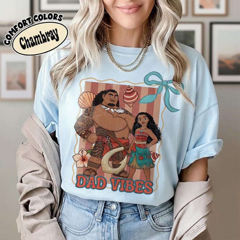 May include: Light blue chambray t-shirt featuring a cartoon illustration of two characters with the text "DAD VIBES". The design includes a large man with tattoos and a young girl, with a seashell and a flower.