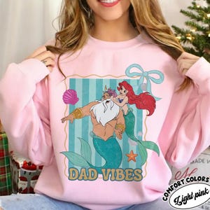May include: Light pink sweatshirt with a graphic of a merman and mermaid with the text "DAD VIBES". The graphic has a blue and gold border with a seashell, bow, and starfish. The sweatshirt is made of Comfort Colors material.