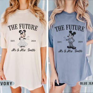 Puede incluir: Dos camisetas con un gr&aacute;fico de Mickey Mouse y Minnie Mouse. El texto en las camisetas dice "The Future Est. 2024 Mr & Mrs. Smith". Una camiseta es de color marfil y la otra es de color azul jean.