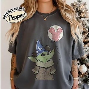 May include: Dark gray t-shirt featuring a cartoon image of Grogu wearing a wizard hat and holding a Mickey Mouse balloon. The shirt has the words "COMFORT COLORS Pepper" in a white oval.