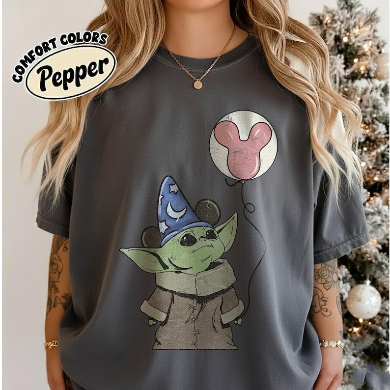May include: Dark gray t-shirt featuring a cartoon image of Grogu wearing a wizard hat and holding a Mickey Mouse balloon. The shirt has the words "COMFORT COLORS Pepper" in a white oval.