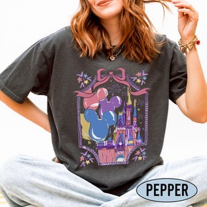 May include: A charcoal gray short-sleeve t-shirt featuring a graphic print of a castle, Mickey Mouse-shaped balloons in blue, purple, and pink, and a pink bow. The design is framed by a decorative border with small flowers and fireworks.