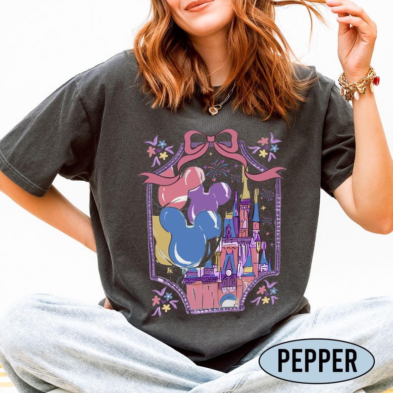 May include: A charcoal gray short-sleeve t-shirt featuring a graphic print of a castle, Mickey Mouse-shaped balloons in blue, purple, and pink, and a pink bow. The design is framed by a decorative border with small flowers and fireworks.