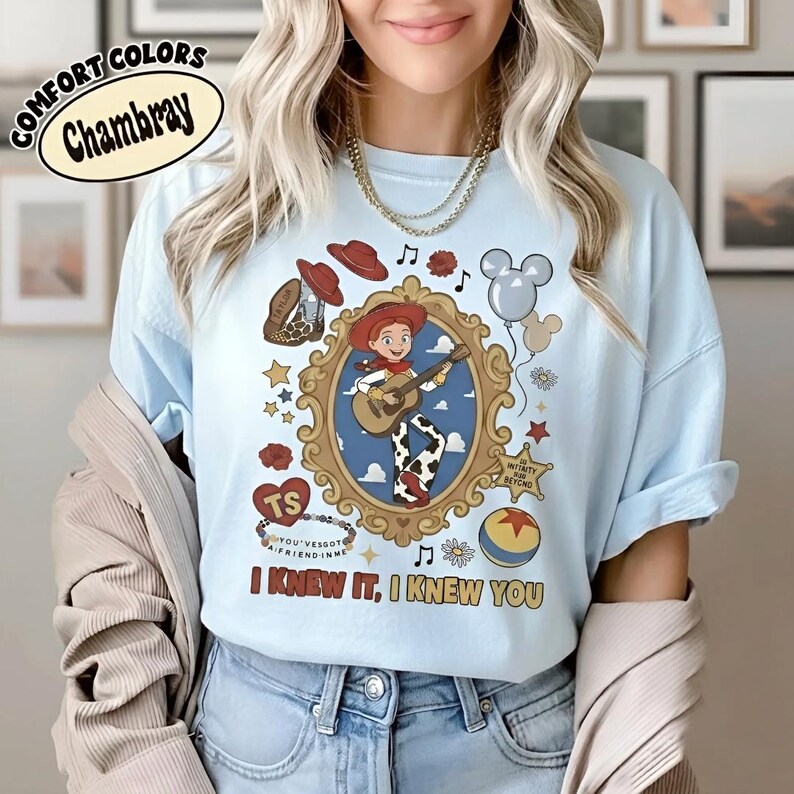 May include: A light blue t-shirt featuring a graphic of a cowgirl playing a guitar inside an ornate gold frame. Surrounding elements include a cowboy hat, boots, balloons, stars, flowers, a ball, and the text "I KNEW IT, I KNEW YOU" at the bottom.