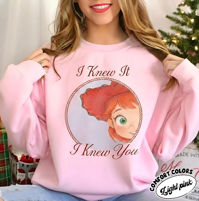 May include: A light pink crewneck sweatshirt featuring a circular graphic of a red-haired character wearing a red hat. The text "I Knew It" appears above the circle and "I Knew You" appears below the circle in a dark brown script font.