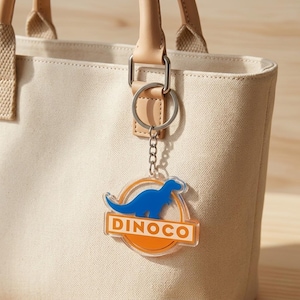 Puede incluir: Un llavero de acr&iacute;lico transparente con la silueta de un dinosaurio azul dentro de un c&iacute;rculo naranja y el texto DINOCO en letras blancas sobre una barra rectangular naranja, sujeto a una anilla de metal plateado en un bolso de lona beige.