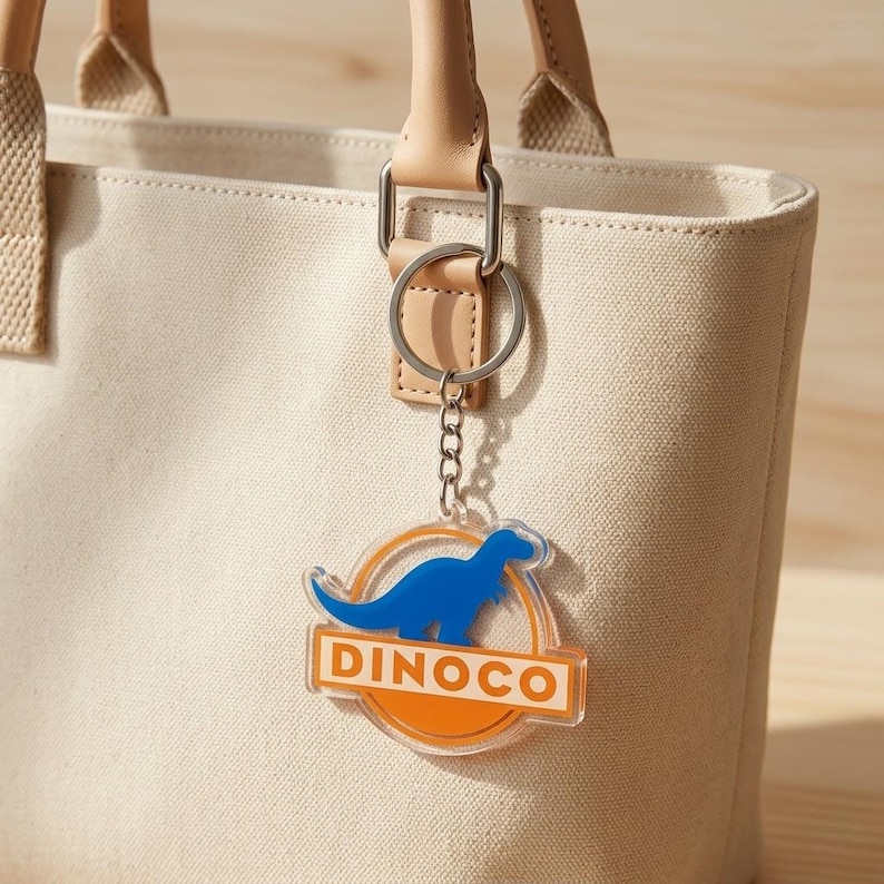 Puede incluir: Un llavero de acr&iacute;lico transparente con la silueta de un dinosaurio azul dentro de un c&iacute;rculo naranja y el texto DINOCO en letras blancas sobre una barra rectangular naranja, sujeto a una anilla de metal plateado en un bolso de lona beige.