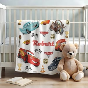 May include: A white fleece blanket featuring a colourful print of animated race cars, including a red car with number 95, a blue car, and a brown tow truck. The design includes the name Robert in red text, checkered flags, trophies, and Route 66 signs.