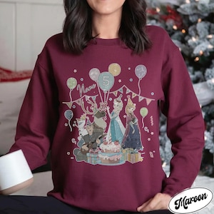 Puede incluir: Sudadera granate con un gr&aacute;fico colorido de personajes de dibujos animados, globos y una tarta de cumplea&ntilde;os. El n&uacute;mero 5 se muestra en un globo, con la palabra "Name" encima de los personajes. La palabra "Maroon" est&aacute; impresa en la parte inferior derecha.