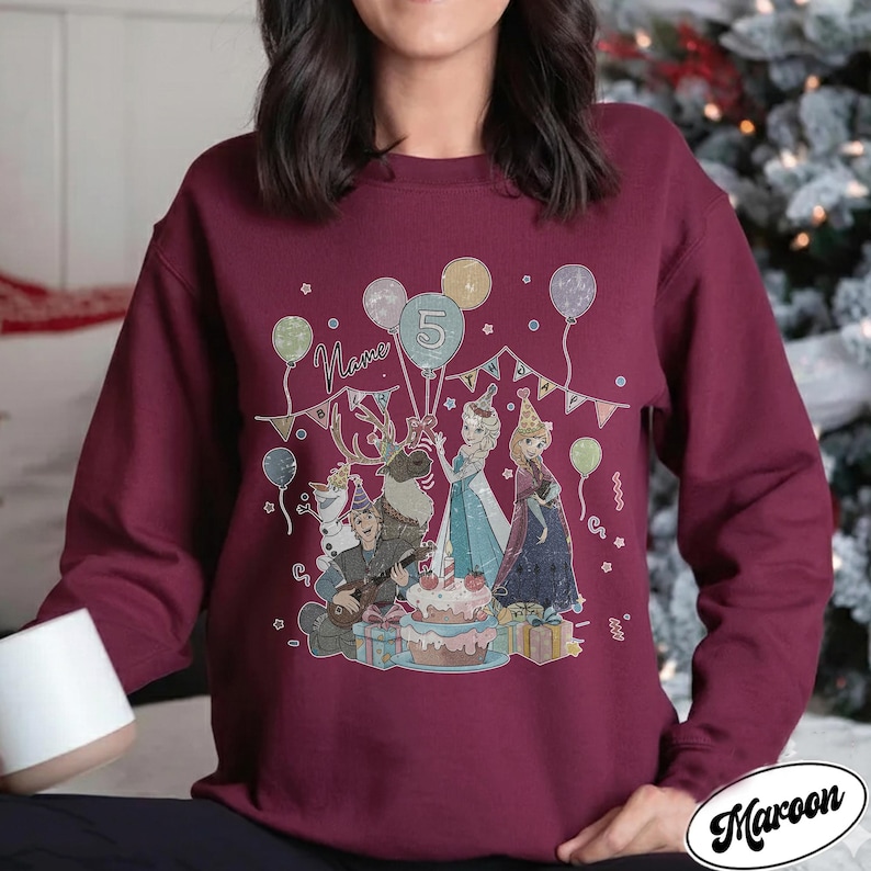 Puede incluir: Sudadera granate con un gr&aacute;fico colorido de personajes de dibujos animados, globos y una tarta de cumplea&ntilde;os. El n&uacute;mero 5 se muestra en un globo, con la palabra "Name" encima de los personajes. La palabra "Maroon" est&aacute; impresa en la parte inferior derecha.