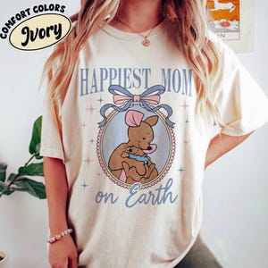 May include: Ivory-colored t-shirt with the text "Happiest Mom on Earth" in light blue. The shirt features a circular graphic of a kangaroo and joey, framed by a pink and blue bow. The words "Comfort Colors" and "Ivory" are in a circle at the top.