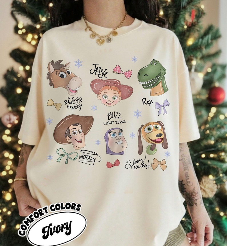 May include: Ivory-colored t-shirt featuring cartoon characters from the Toy Story franchise. The design includes images of Woody, Buzz Lightyear, Jessie, Rex, and others, with their names and decorative bows. The shirt also has the words "COMFORT COLORS" and "Ivory".