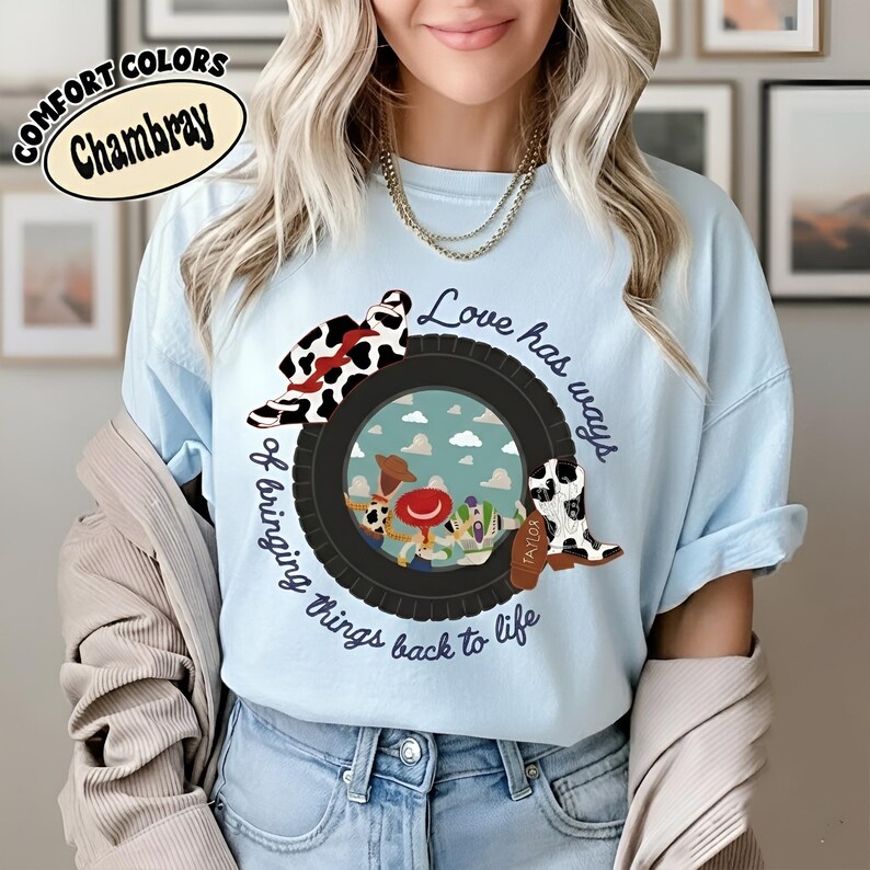 Puede incluir: Una camiseta azul claro con un gr&aacute;fico de un neum&aacute;tico negro que rodea un fondo azul cielo con personajes de dibujos animados. El dise&ntilde;o incluye un sombrero con estampado de vaca, una bota con estampado de vaca y el texto: Love has ways of bringing things back to life.
