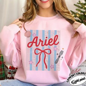 May include: Light pink sweatshirt with the name "Ariel" in red script. The design includes a red bow, stars, a fork, and a seashell on a striped background of blue and pink. The sweatshirt has the words "Comfort Colors" and "Light pink" in a circular logo.