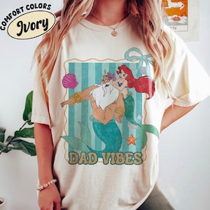 May include: Ivory-colored t-shirt with a cartoon graphic featuring a merman, Ariel, and the text "DAD VIBES." The graphic is framed with a blue and white striped background. The shirt also has the words "COMFORT COLORS" and "Ivory" printed on it.