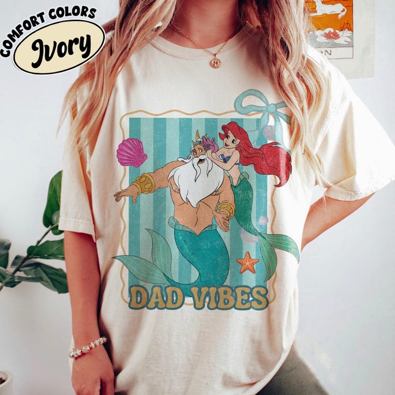 May include: Ivory-colored t-shirt with a cartoon graphic featuring a merman, Ariel, and the text "DAD VIBES." The graphic is framed with a blue and white striped background. The shirt also has the words "COMFORT COLORS" and "Ivory" printed on it.