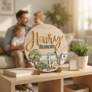 May include: A circular wooden sign featuring the name Henry in large yellow script and Robert in smaller dark green text. The base displays a layered scene with trees, a white fence, a small honey pot, and figures of a bear and a small animal.