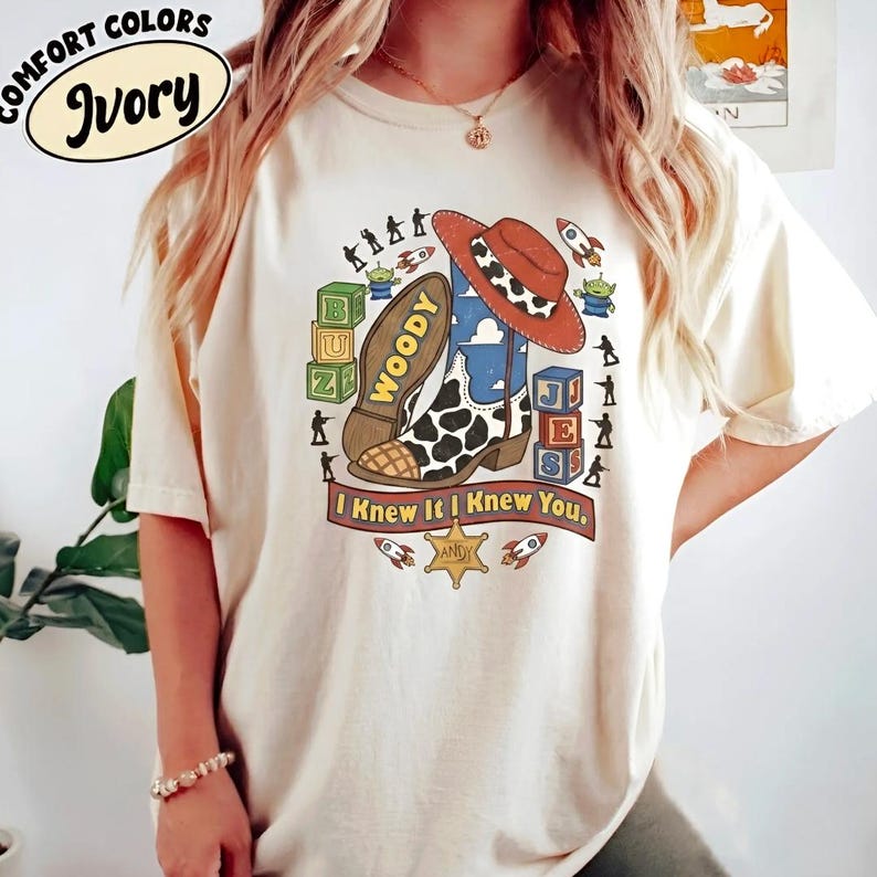 May include: An ivory short-sleeve t-shirt featuring a graphic print of a cowboy boot, a hat, alphabet blocks, and small toy figures. The text reads "WOODY", "BUZZ", "JES", "I KNEW IT I KNEW YOU", and "ANDY" on a sheriff star badge.