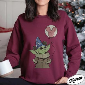 May include: Maroon sweatshirt featuring a cartoon character with large ears, wearing a wizard hat with a crescent moon and stars. The character is holding a balloon with a Mickey Mouse head design. The word "Maroon" is printed on the bottom right.