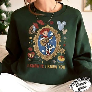 May include: A dark green crewneck sweatshirt featuring a graphic print of a cowgirl character playing a guitar inside an ornate gold frame. The design includes a cowboy hat, balloons, a star badge, a ball, and the text "I KNEW IT, I KNEW YOU".