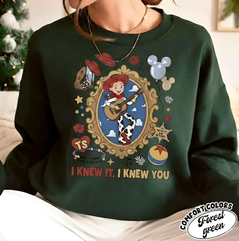 May include: A dark green crewneck sweatshirt featuring a graphic print of a cowgirl character playing a guitar inside an ornate gold frame. The design includes a cowboy hat, balloons, a star badge, a ball, and the text "I KNEW IT, I KNEW YOU".