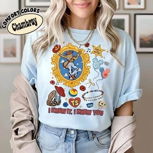 Puede incluir: Una camiseta azul claro con un estampado gr&aacute;fico que incluye un marco dorado ornamentado, un sombrero de vaquero, una guitarra, una estrella con el texto INFINITY AND BEYOND, globos, una pulsera de cuentas con el texto YOU'VE GOT A FRIEND IN ME y el texto I KNEW IT, I KNEW YOU.