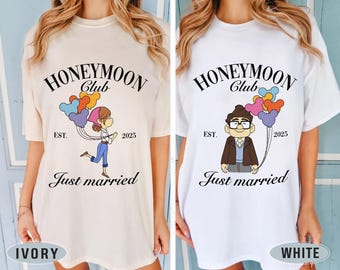May include: Two white t-shirts with a graphic design of a cartoon couple holding balloons that say "Honeymoon Club" with the text "Just married" and the year "2025". The t-shirts are shown on models wearing the shirts. The t-shirt on the left is ivory and the t-shirt on the right is white.
