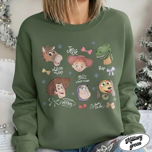 May include: Military green sweatshirt featuring cartoon character portraits from the movie Toy Story. The design includes images of Woody, Buzz Lightyear, Jessie, Rex, and others, with their names and decorative elements.