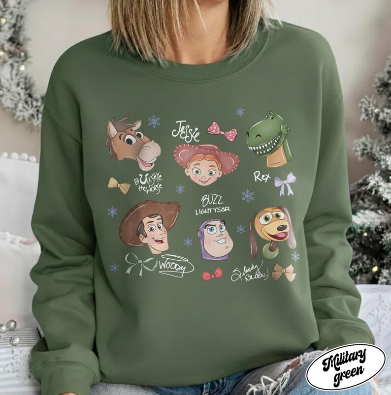 May include: Military green sweatshirt featuring cartoon character portraits from the movie Toy Story. The design includes images of Woody, Buzz Lightyear, Jessie, Rex, and others, with their names and decorative elements.