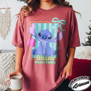 May include: Crimson Comfort Colors t-shirt featuring a cartoon illustration of Stitch from Lilo & Stitch. The design includes Stitch, a seashell, and the text "Ohana Means Family" in yellow. The shirt has short sleeves.