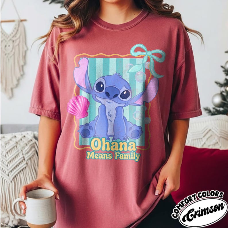 May include: Crimson Comfort Colors t-shirt featuring a cartoon illustration of Stitch from Lilo & Stitch. The design includes Stitch, a seashell, and the text "Ohana Means Family" in yellow. The shirt has short sleeves.