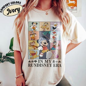 May include: Ivory-colored t-shirt featuring a colorful graphic of various cartoon characters in a grid pattern. The graphic includes Mickey Mouse, Donald Duck, and Winnie the Pooh. Text on the shirt reads "IN MY RUNDISNEY ERA."