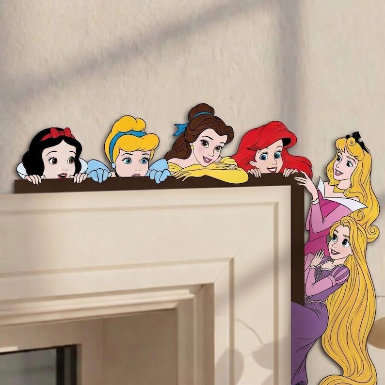 May include: A set of decorative wall decals featuring six animated princesses. Snow White, Cinderella, Belle, Ariel, Aurora, and Rapunzel are depicted peeking over the top edge of a door frame or rectangular structure with colorful outfits.