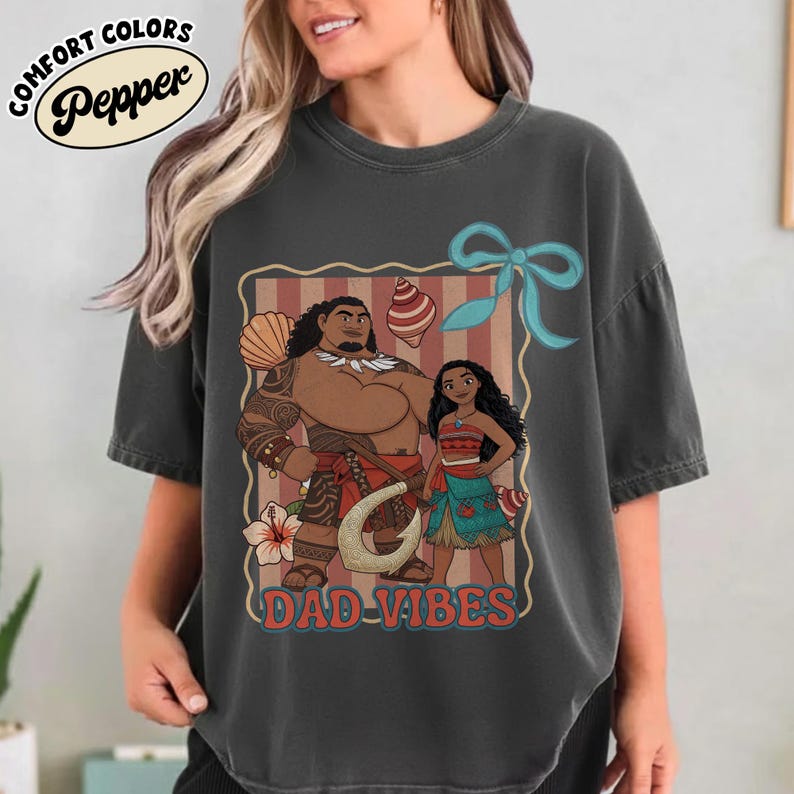 May include: Dark gray t-shirt featuring a cartoon illustration of two characters with the text "DAD-VIBES". The design includes a bow, seashell, and flower accents. The shirt has the words "COMFORT COLORS" and "Pepper" in a circle.