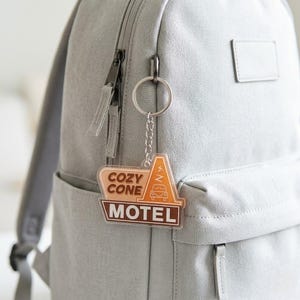 Puede incluir: Un llavero de acr&iacute;lico transparente con un gr&aacute;fico de un edificio en forma de cono de tr&aacute;fico, el contorno de un coche y el texto COZY CONE MOTEL en tonos marr&oacute;n y naranja, unido a una anilla y cadena de metal plateado en una mochila gris.