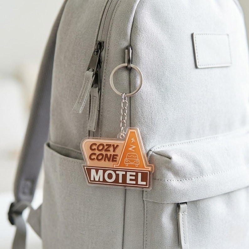 Puede incluir: Un llavero de acr&iacute;lico transparente con un gr&aacute;fico de un edificio en forma de cono de tr&aacute;fico, el contorno de un coche y el texto COZY CONE MOTEL en tonos marr&oacute;n y naranja, unido a una anilla y cadena de metal plateado en una mochila gris.