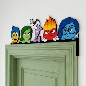 May include: A black door topper featuring five colorful cartoon characters from Inside Out: Joy, Disgust, Fear, Anger, and Sadness. The characters are arranged in a row, each with distinct facial expressions and colors, mounted on a black frame.