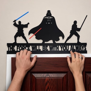 May include: A black metal wall art piece featuring silhouettes of three figures, including a central character in a cape and two figures holding lightsabers. The blue and red lightsabers are colored. The text reads THE FORCE WILL BE WITH YOU ALWAYS.