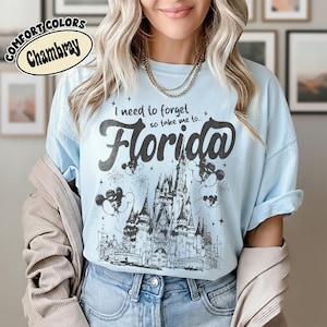 May include: Pale blue t-shirt with the words "I need to forget so take me to... Florida" in black script. The graphic features a detailed illustration of a castle, fireworks, and Mickey Mouse balloons. The shirt is labelled "Comfort Colors Chambray".