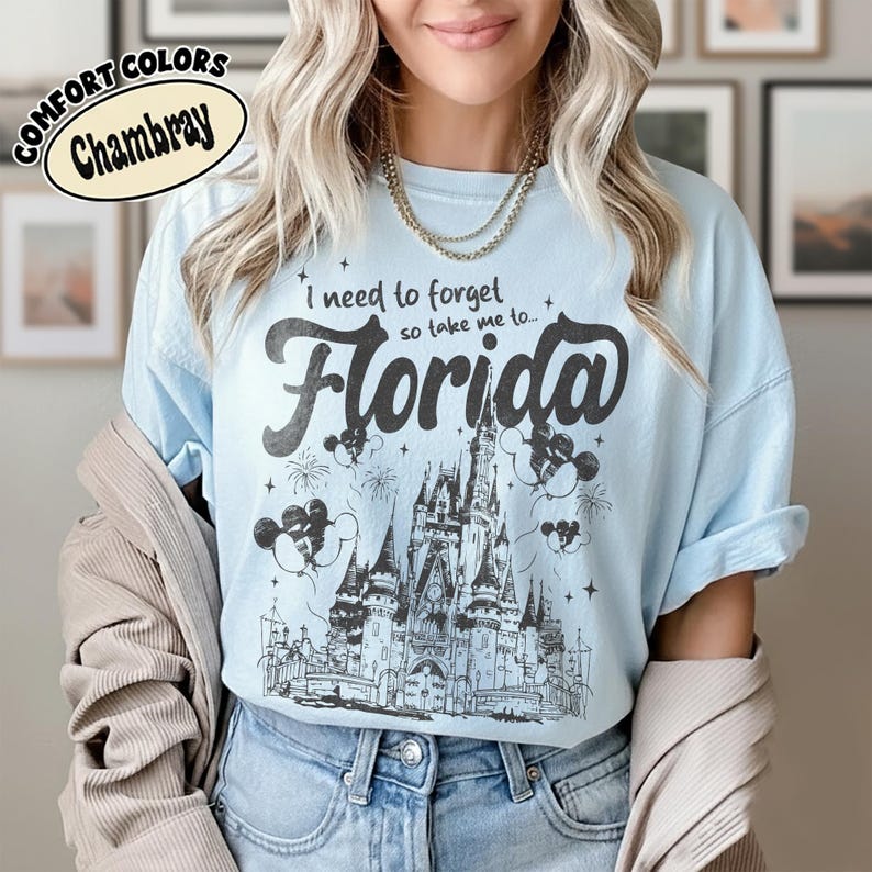 May include: Pale blue t-shirt with the words "I need to forget so take me to... Florida" in black script. The graphic features a detailed illustration of a castle, fireworks, and Mickey Mouse balloons. The shirt is labelled "Comfort Colors Chambray".