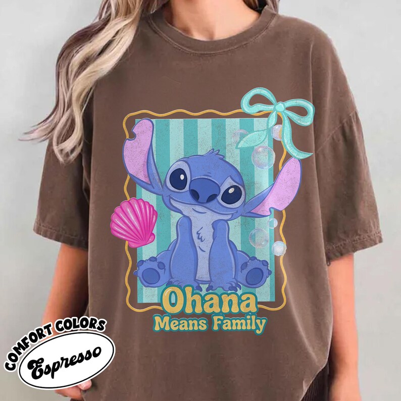 May include: Espresso-colored t-shirt featuring a blue cartoon character with large ears, inside a teal and yellow frame. The text "Ohana Means Family" is below the character. A pink seashell and a blue bow are also present.